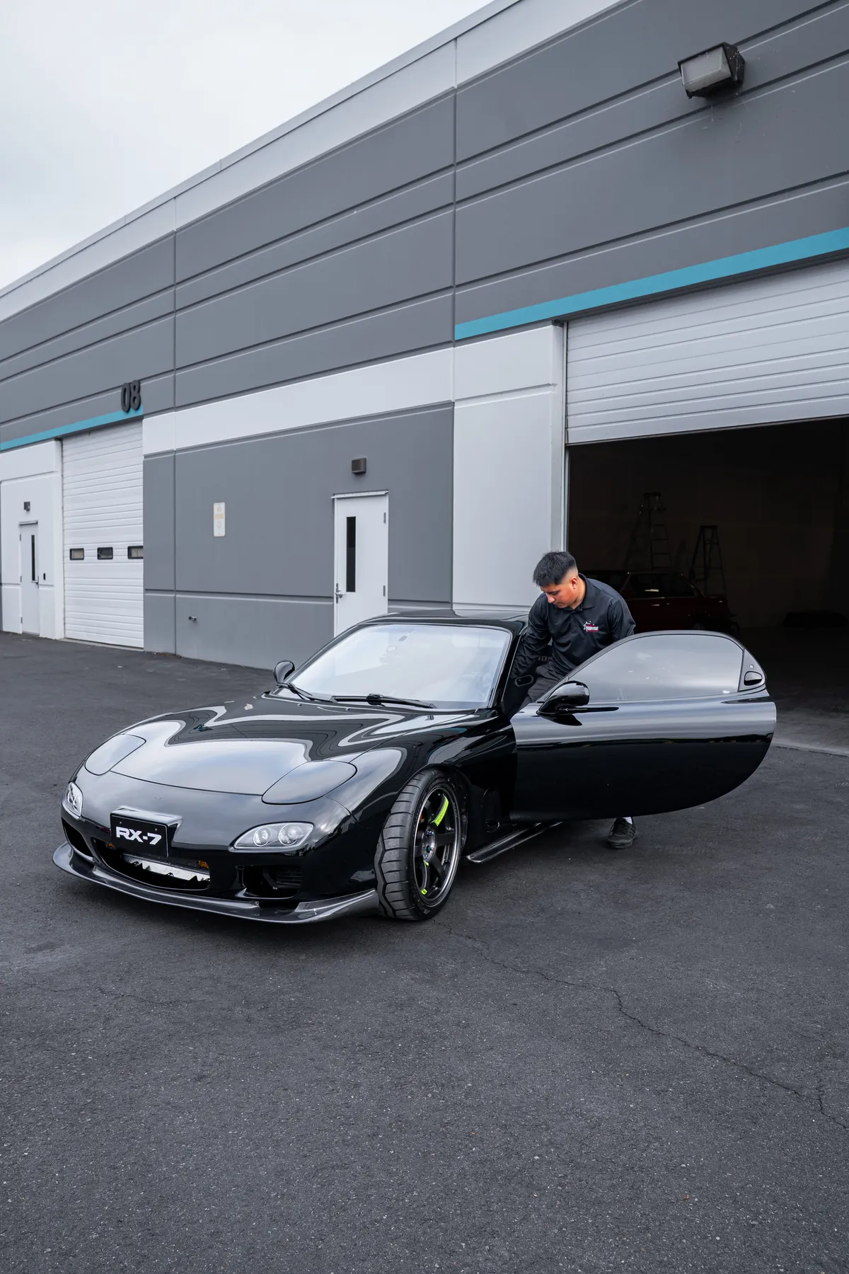 A technician opening the door of a black Mazda RX7 outside the studio